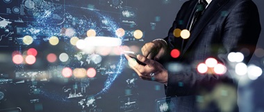 A futuristic image shows man in a business suit holding a smartphone while in the background there are futuristic light trails and examples of data being projected using holographic technology 