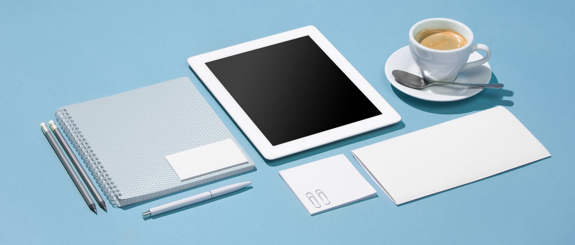 The essential tools of a mobile or freelance office worker are laid out neatly across a pale blue desk: A paper pad, tablet computer, espresso coffee cup, pens and note pads.
